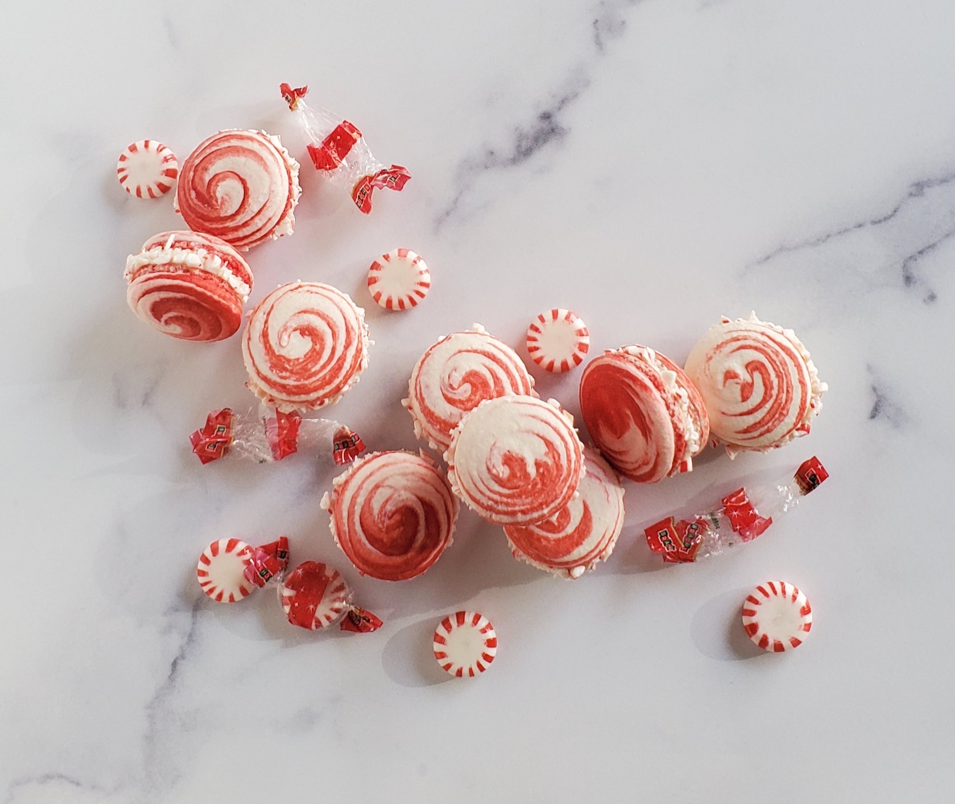 Peppermint swirl macarons in the shape of a "C" scattered with peppermint candies and their wrappers on a white marble surface.