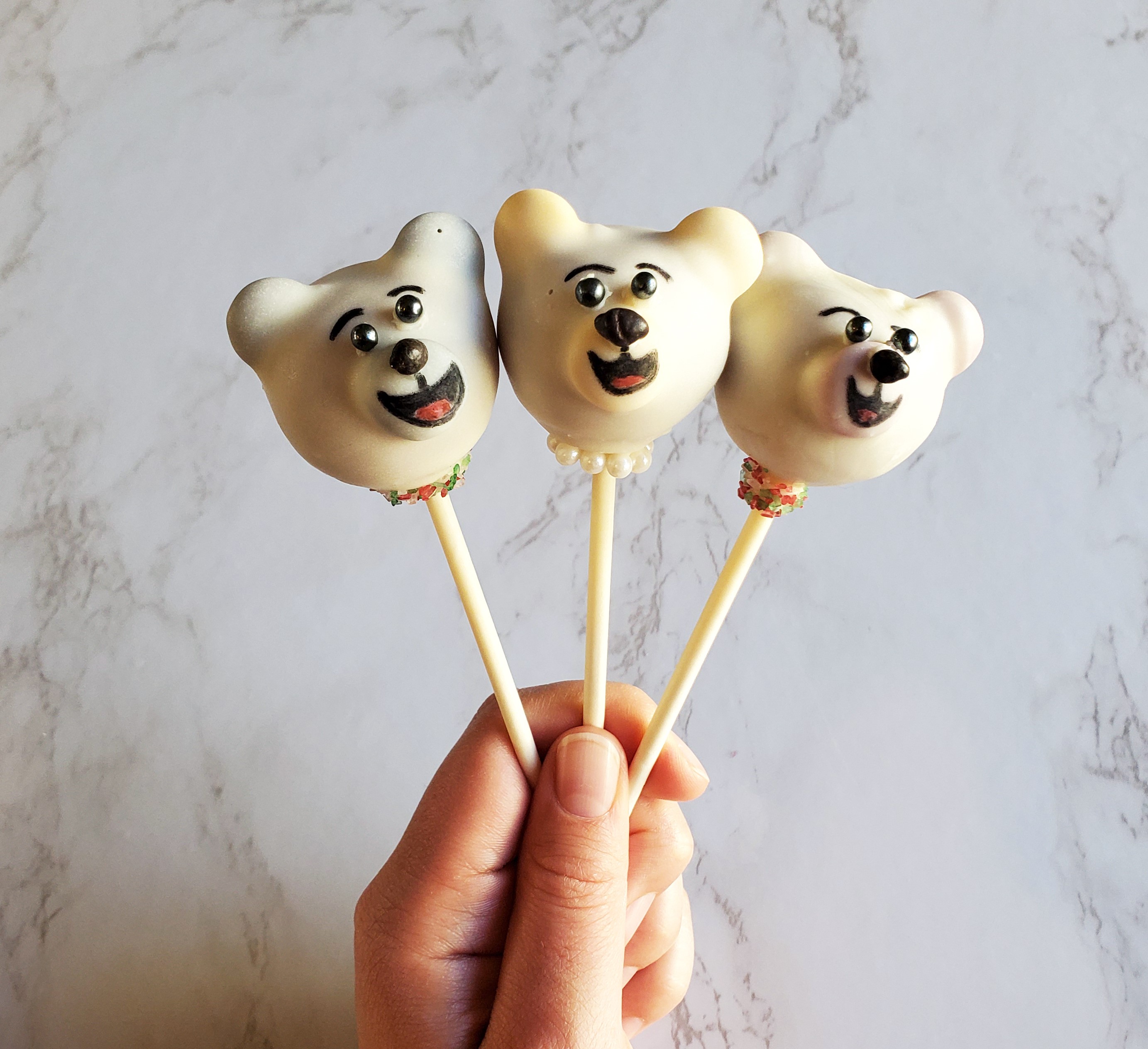 Three Polar Bear cake pops in someone's left hand, in front of a white marbled background. The polar bear face have two black pearl sprinkle eyes, a black candy chip nose and a happy expression drawn on with a food safe marker.