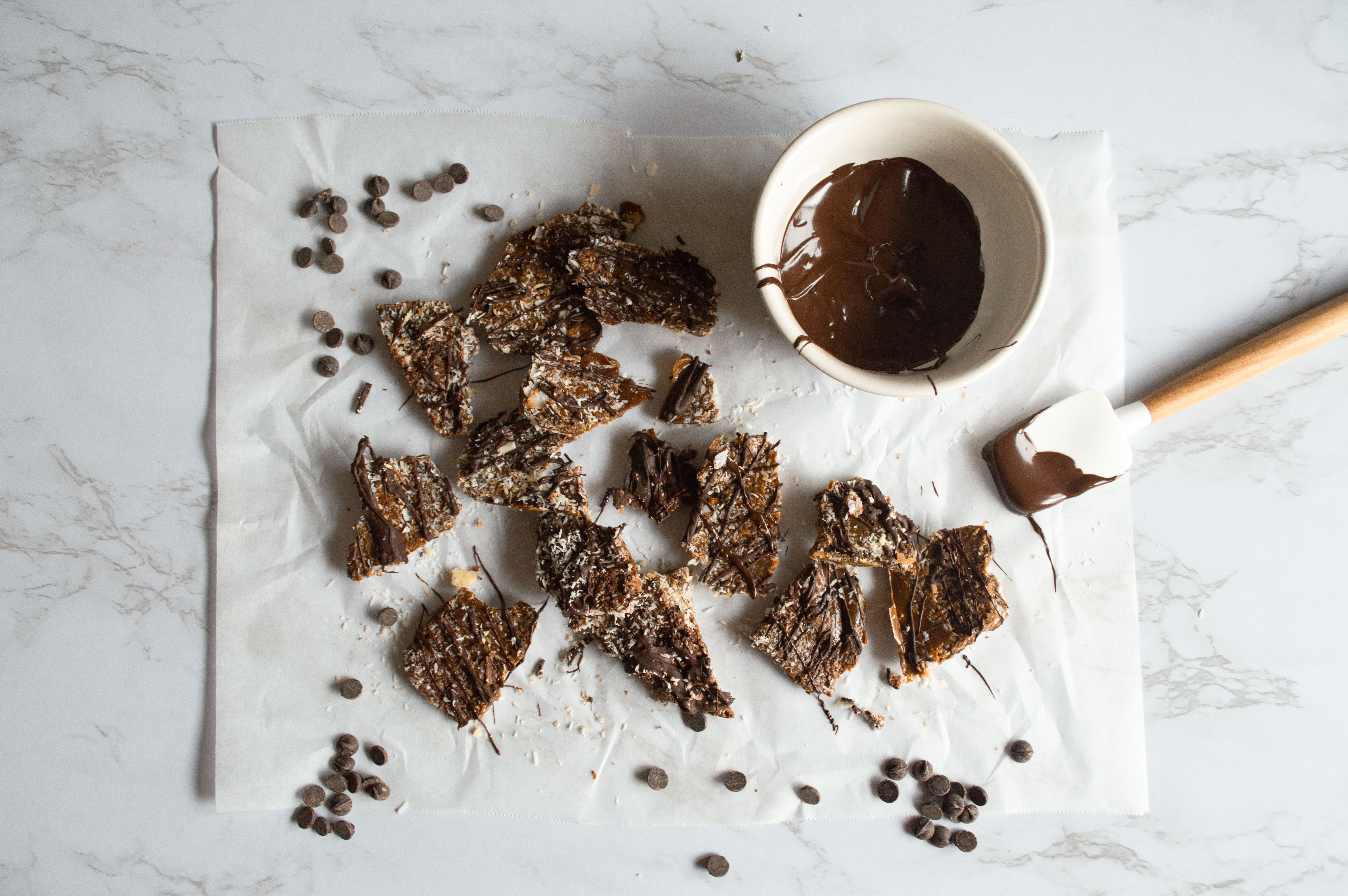 On a white marble countertop, there is a rectangular piece of parchment paper. On top of it, there are pieces of Samoas Vegan Toffee. There are chocolate chips, pieces of coconut and in the upper righthand corner, a small white bowl of melted chocolate and a small spatula lying on the parchment paper with melty chocolate on the tip.