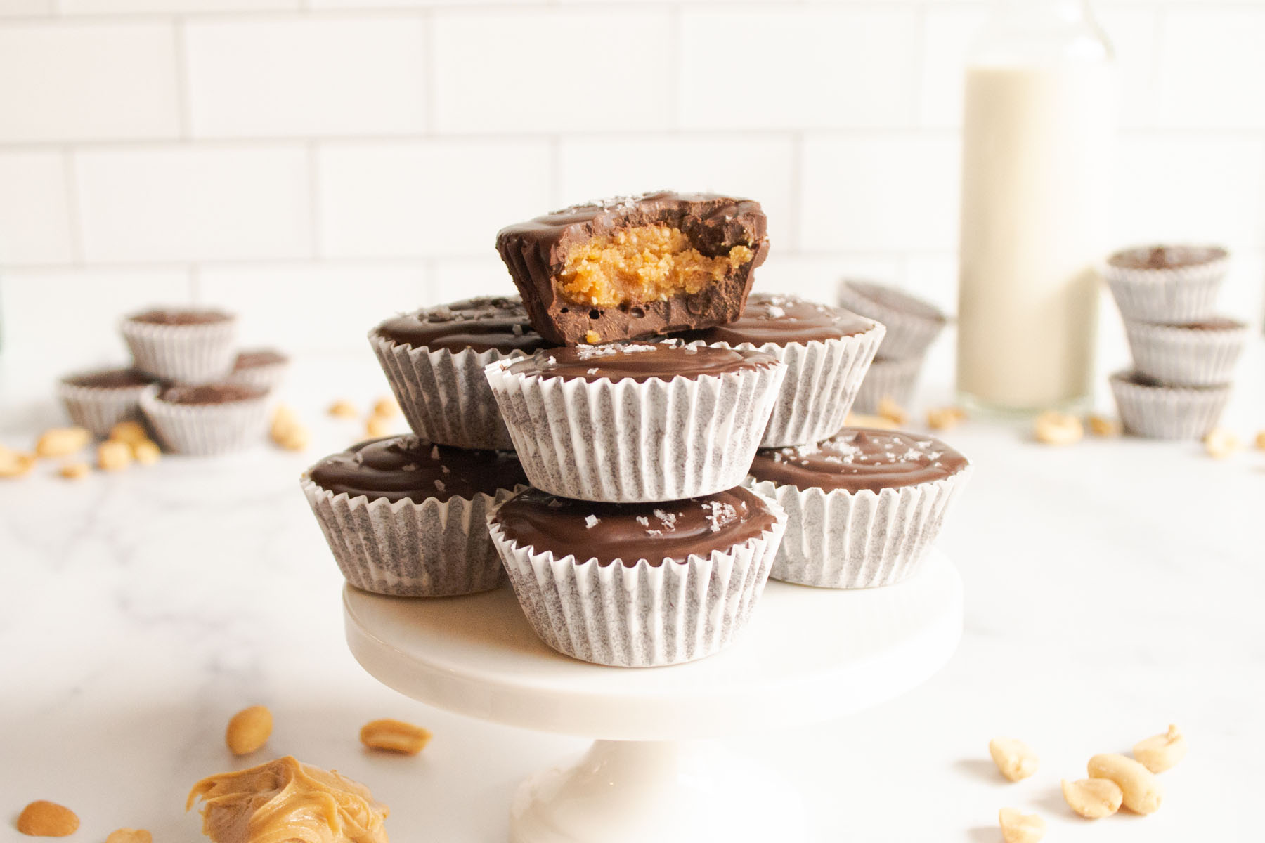 On a short cupcake stand there are homemade peanut butter cups stacked. The one on top has a large bite out of it. In the foreground we see peanuts and a spoonful of peanut butter. In the background we see a glass of milk, more peanuts and peanut butter cups.