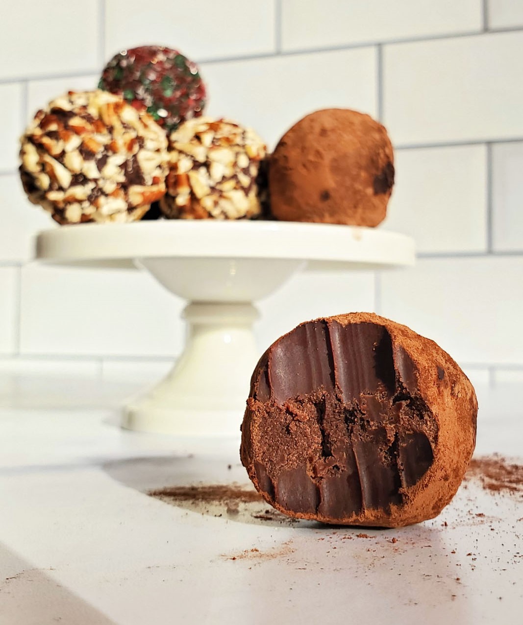 A truffle covered in cocoa powder, with a bite out of it sits in the foreground. In the background, on a cake pedestal sits five more truffles covered in various toppings including, chopped pecans, holiday red/green sprinkles and another truffle covered in cocoa powder. Behind the truffles is a subway tile wall.