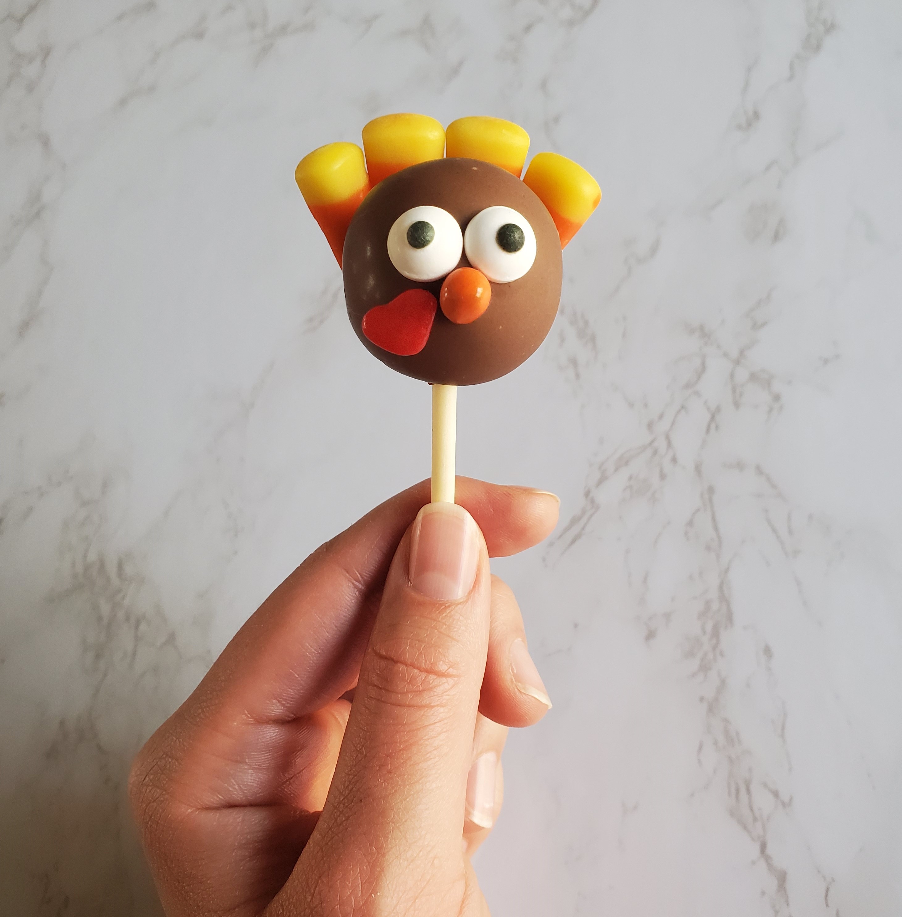 Turkey cake pop with candy eyeballs, orange candy chip beaks, cinnamon heart gobbles and candy corn feathers in a hand against a white marble wall.