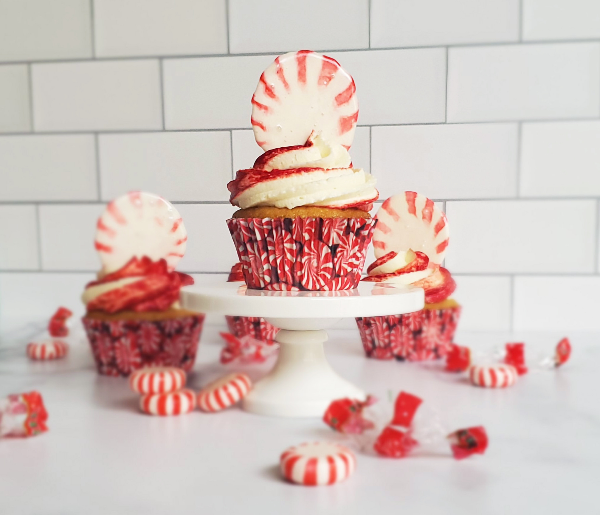 Vanilla Peppermint cupcakes with a melted peppermint candy piece on top. The center cupcake is on a mini pedestal and surrounded by peppermint candies and their wrappers.