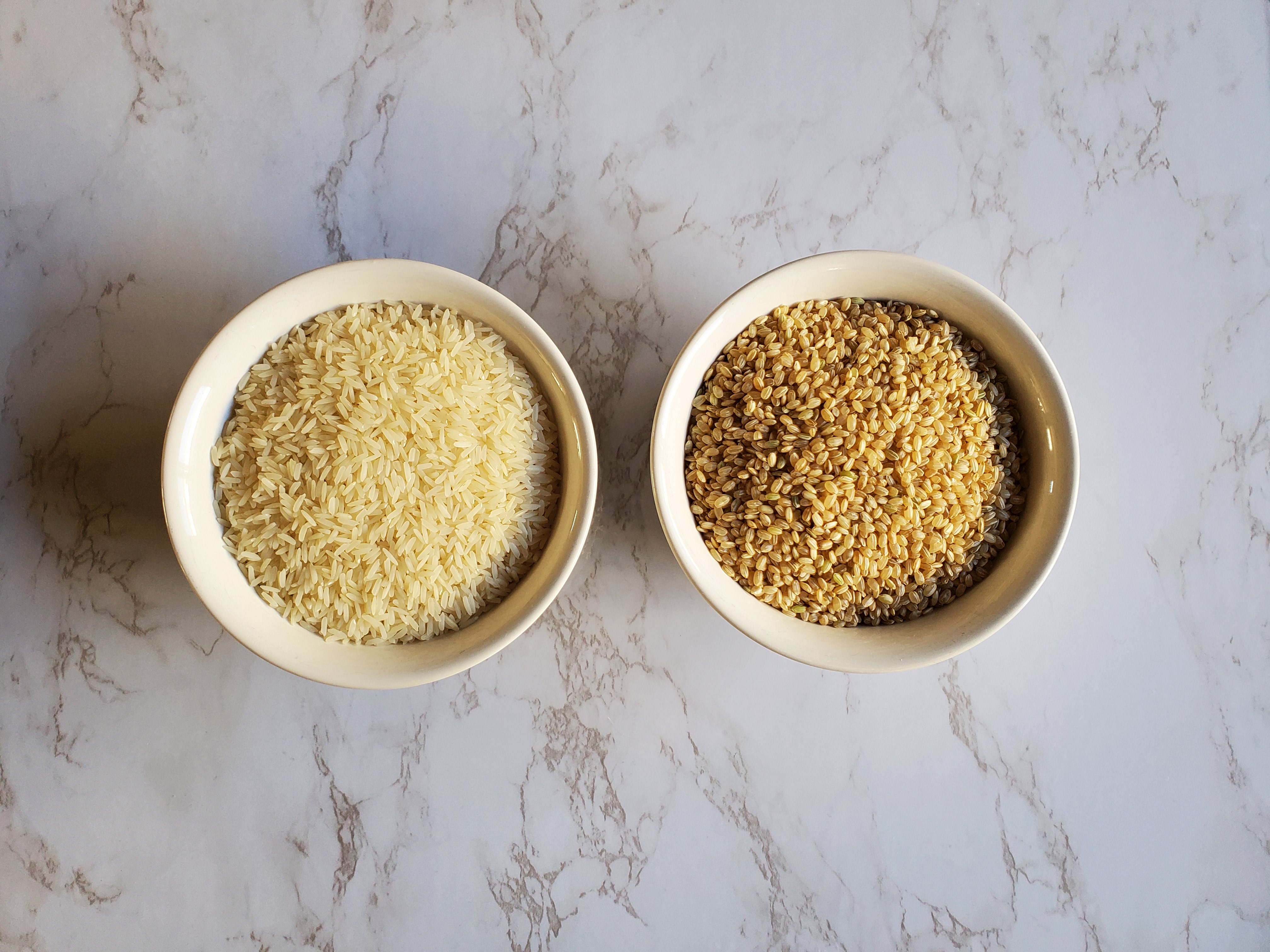 A white bowl on the left is filled with white jasmine rice and the white bowl on the left if filled with brown short grain rice, both against a white marble background.