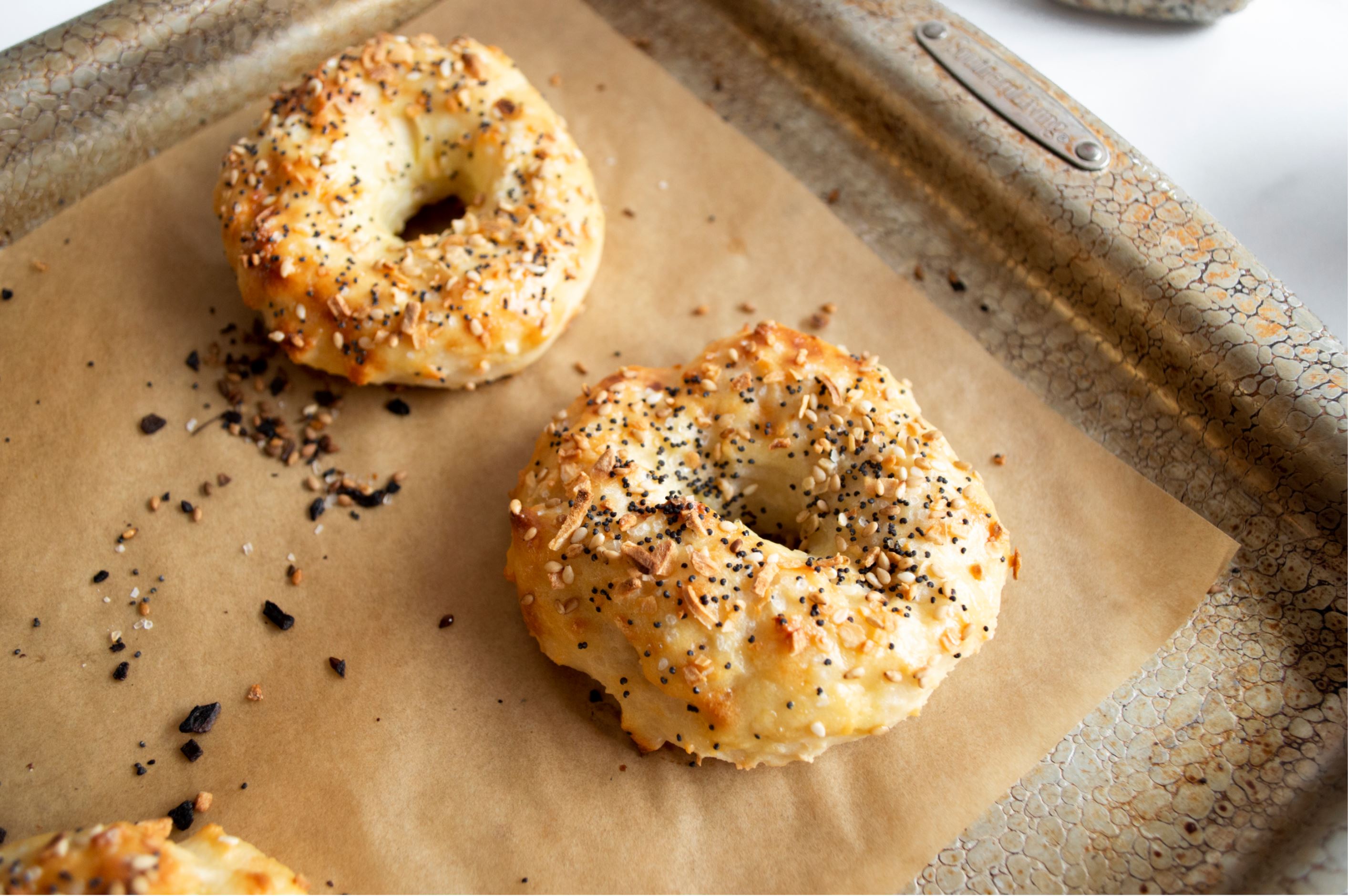 Two Greek Yogurt Everything Bagels sit on brown parchment paper on a metal pan. The parchment has pieces of everything bagel seasoning scattered on it.