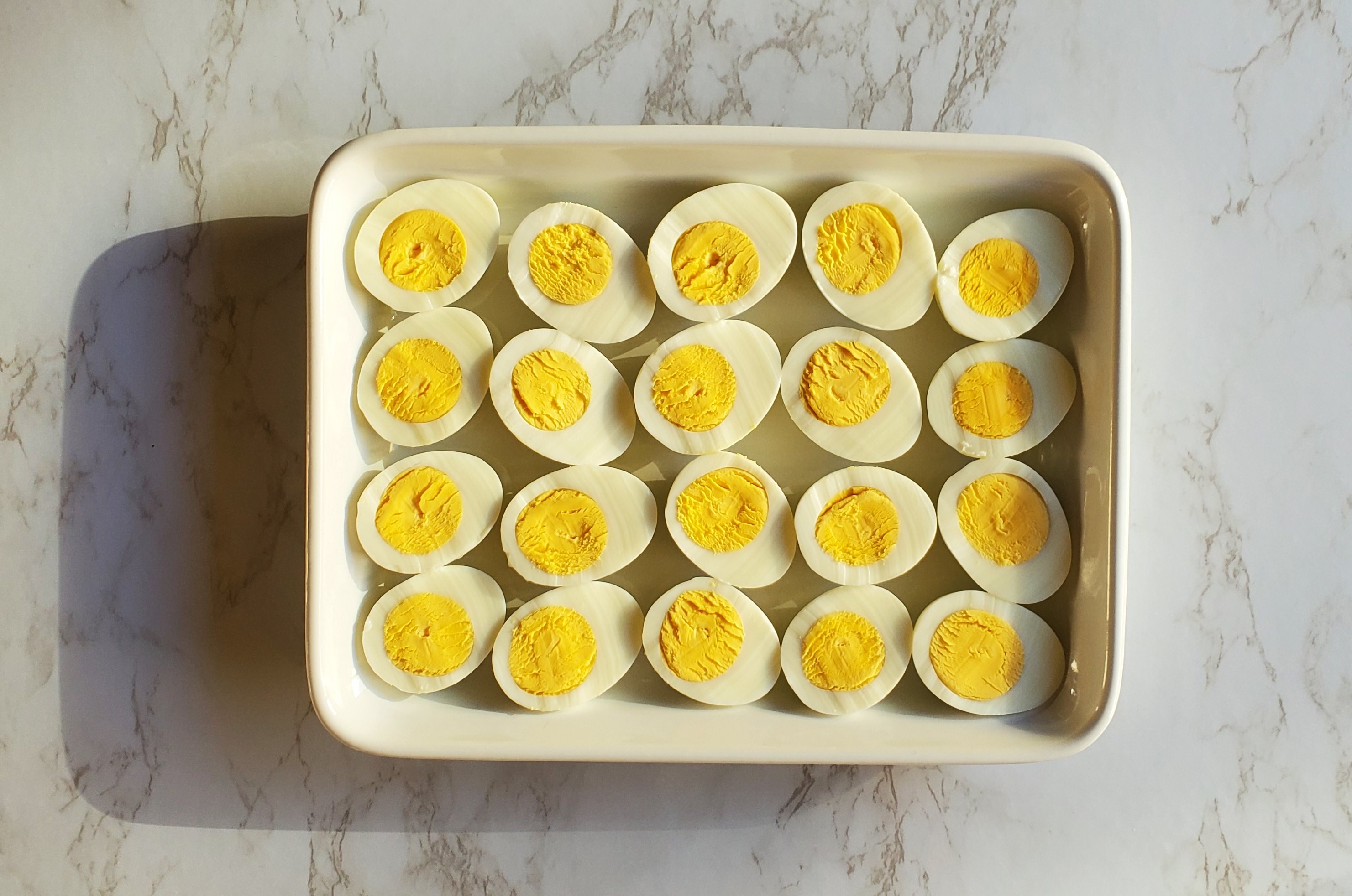 A white rectangular ceramic platter on a marble counter top filled with halved hard boiled eggs.