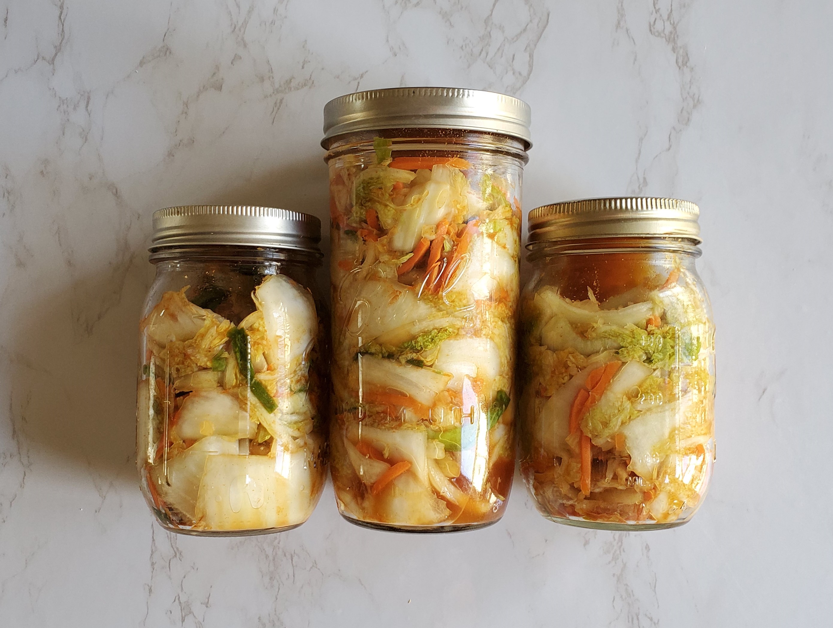 Three jars of kimchi laying on a white marble background in Mason Ball jars.