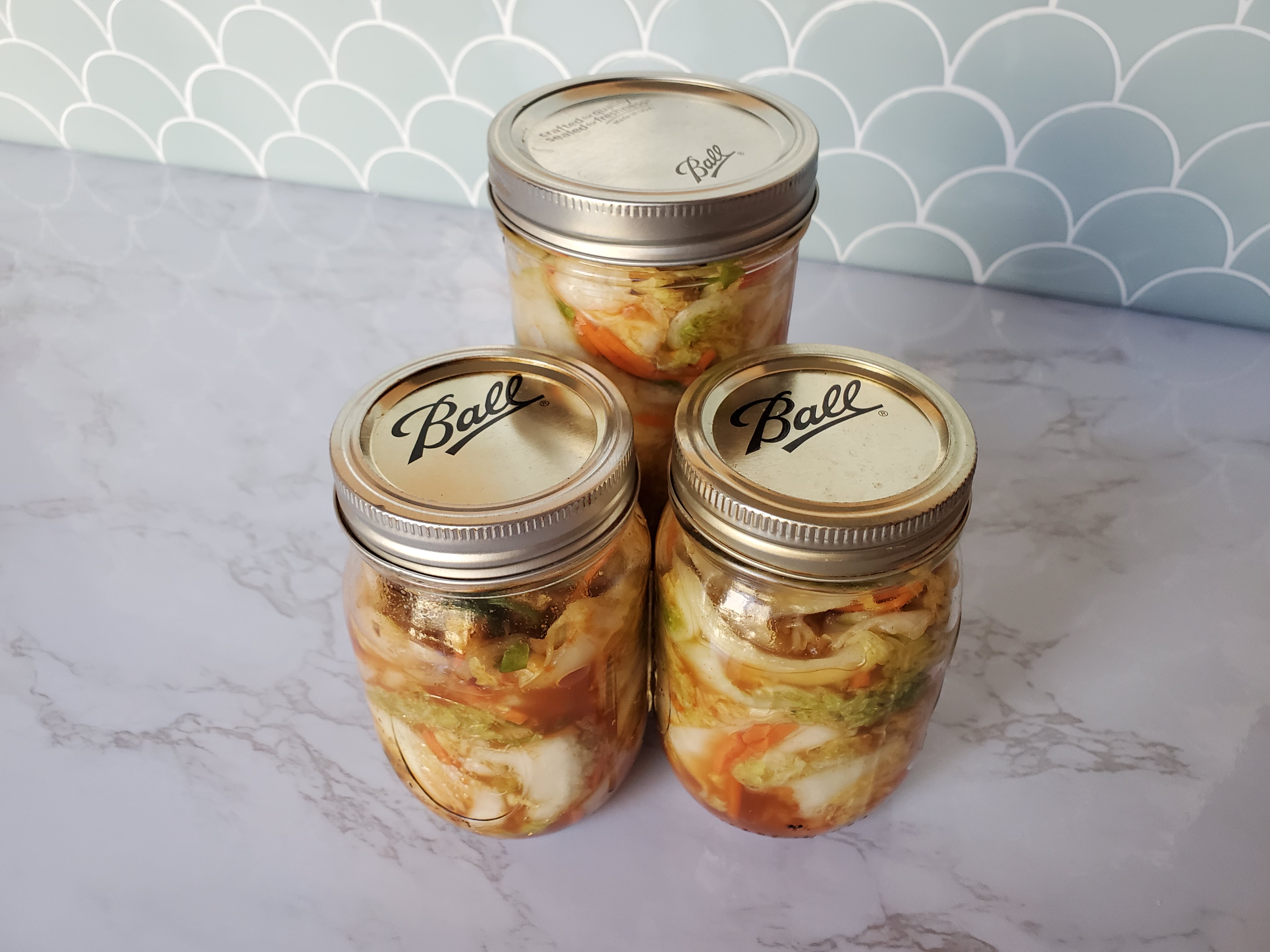 Kimchi in Ball jars on a granite countertop and turquoise shell tile backsplash.