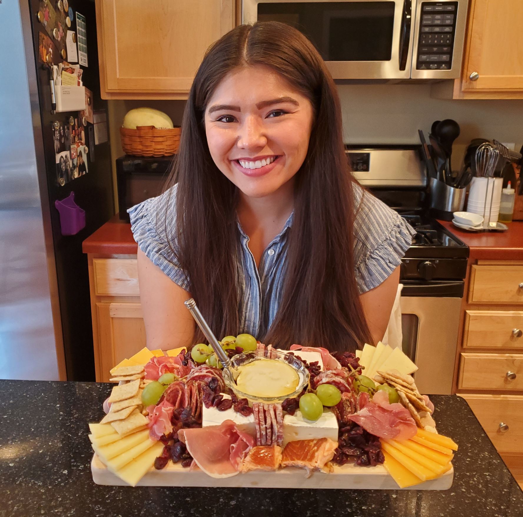 A picture of me, a smiling brunette, hair down. I am holding a charcuterie board. It is filled with prosciutto, salami, smoked salmon, goat cheese drizzled with honey, brie, craisins, grapes, gounda, merlot cheese, crackers and marcona almonds. In the background is my kitchen, oven, microwave and fridge. You can see kitchen utensils in a cup on the counter.