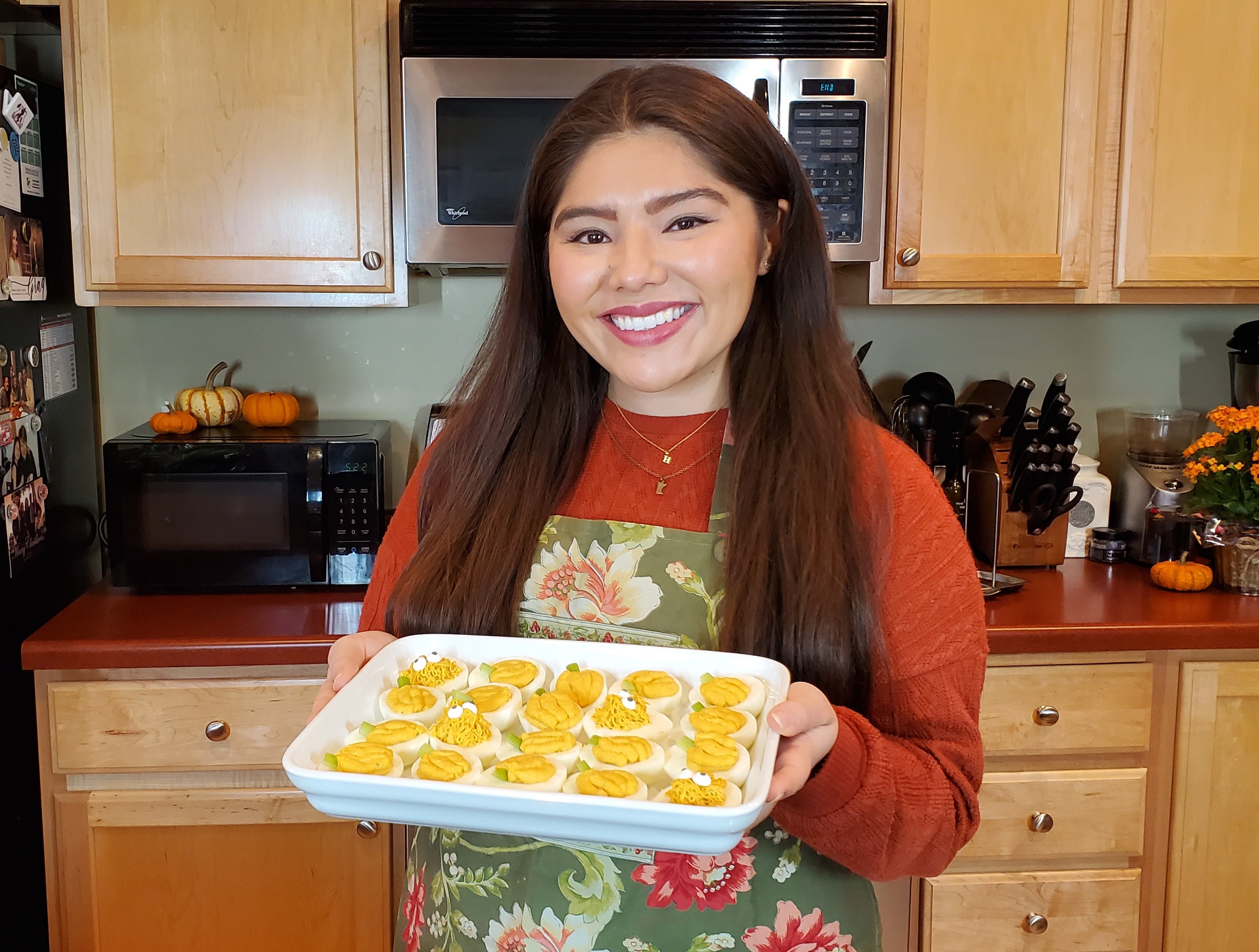 Lauren Hugh of hugh eats with you holding a white rectangular platter filled with hard boiled eggs in her kitchen. She wear a burnt orange knit sweater and a green and pink floral fall apron.