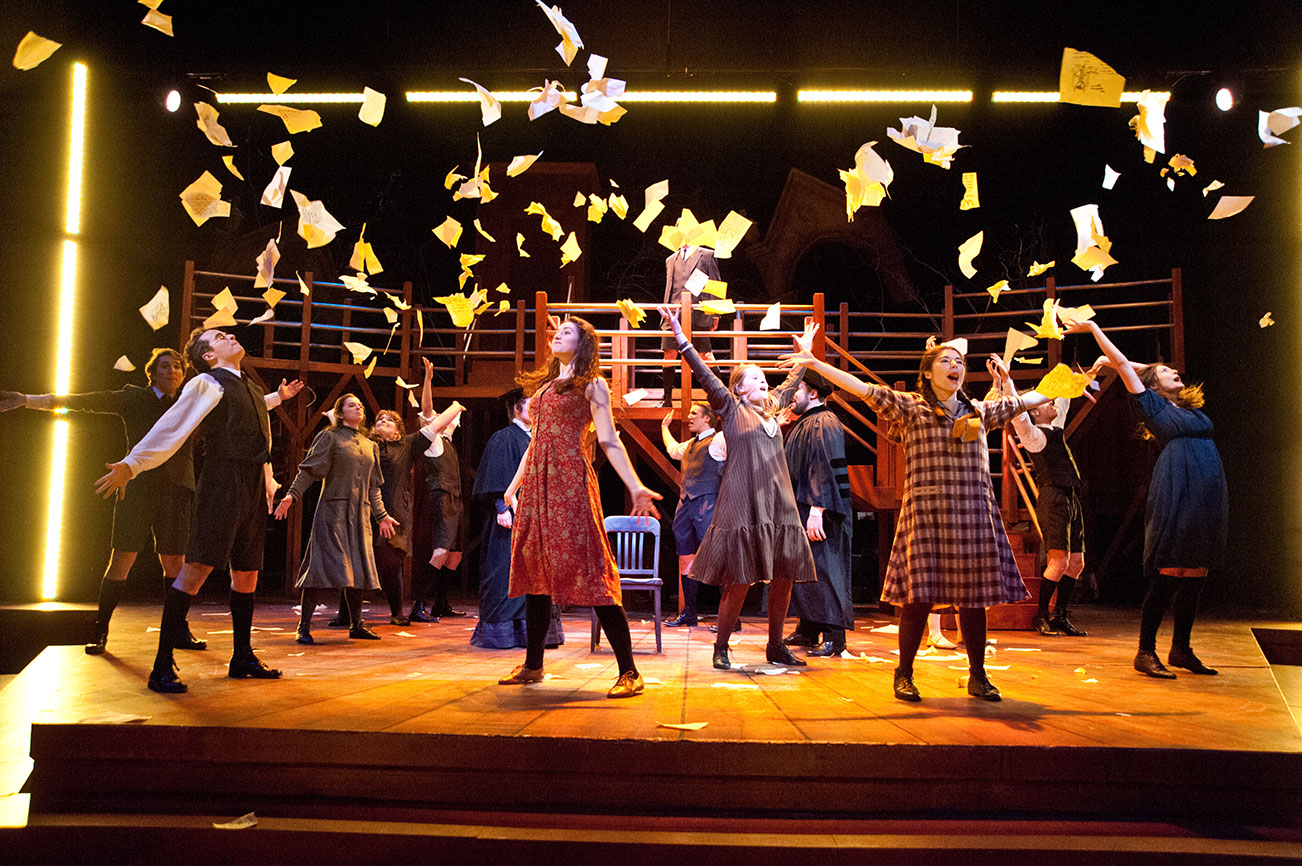 A large group of 19th century school children throw their school papers into the air on a stage. There is a singular chair in the middle, a professor (punishing the students) at each side. The stage is lit with yellow LED lights. There is a rustic wooden stage, ripped papers spread across. And a wooded, raised, bridge-like upper-deck.