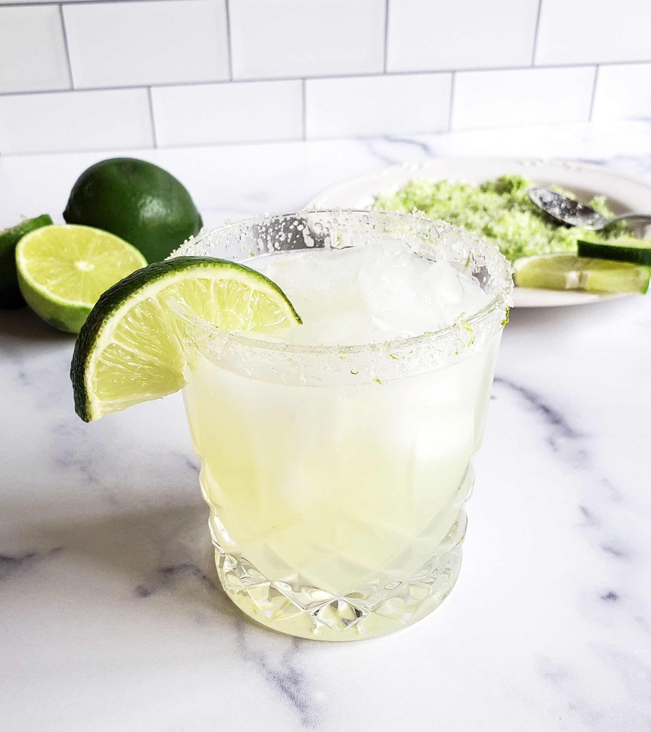 A margarita garnished with a lime wedge sits in front of cut up limes and a plant of a sugar/salt mixture.